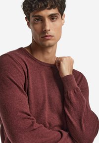 Burgunderfarbener Strickpullover mit Rundhalsausschnitt, langen Ärmeln und strukturiertem Stoff, präsentiert an einem Modell mit kurzen lockigen Haaren vor einem einfarbigen Hintergrund.
