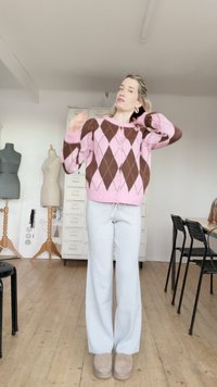 Roze en bruine argyle vest met knoopdetails, gedragen over lichtgrijze joggingbroek en beige laarzen; houten vloer en kledingrekken op de achtergrond.