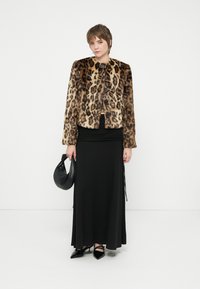 Persona vestida con una chaqueta de piel de leopardo, una falda larga negra, tacones negros de punta y sosteniendo un bolso negro sobre un fondo blanco.