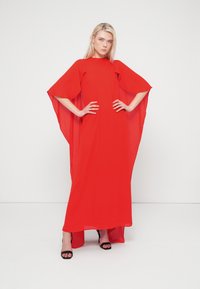 Κόκκινο μακρύ φόρεμα με χαλαρή, ρέουσα γραμμή και φαρδιά μανίκια. Κατασκευασμένο από ελαφρύ ύφασμα με ψηλή σκίσιμο. Συνδυάζεται με μαύρα τακούνια.