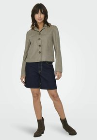 Brun mockakavaj med krage och fem knappar, matchad med mörka denimshorts med gula sömmar. Modellen bär bruna ankelstövlar.