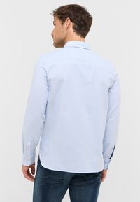 Jasnoniebieska, w paski, koszula z długim rękawem, z kołnierzykiem zapinanym na guziki, jedynym guzikiem przy mankietach oraz zaokrąglonym dołem; materiał wydaje się gładki i przewiewny.