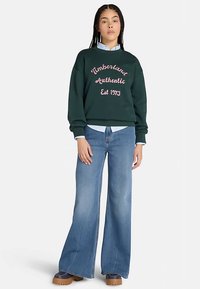 Sudadera verde con texto rosa bordado, combinada con vaqueros acampanados de lavado claro. La modelo lleva una camisa con cuello debajo. Calzado voluminoso.