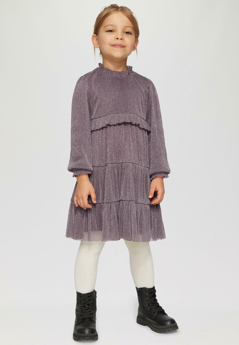 Robe violette à plusieurs niveaux, avec des manches longues, un ourlet volanté et un tissu texturé. Portée avec des collants blancs et des bottes noires à lacets.