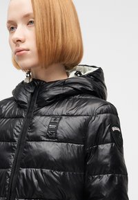 Νεαρό άτομο με ίσια κόκκινα μαλλιά, ντυμένο με μαύρο γυαλιστερό μπουφάν τύπου puffer με κουκούλα και μπαλώματα με το λογότυπο Blauer U.S.A. στο στήθος και το μανίκι.