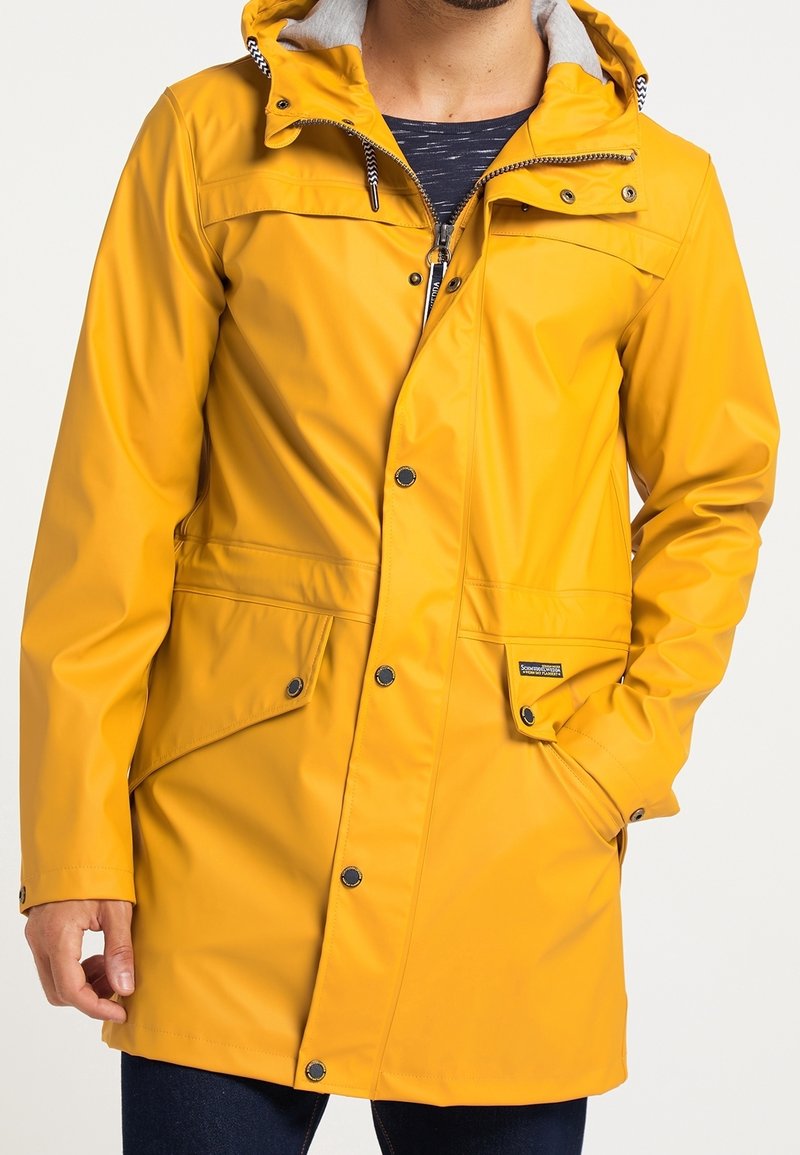 Homme portant une veste imperméable jaune vif avec capuche, fermeture éclair avant, boutons-pression et poches à rabat, main dans la poche.