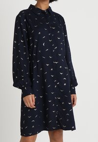 Abito lungo a maniche lunghe blu navy con piccoli stampati di uccelli bianchi, chiusura frontale con bottoni, polsini arricciati e colletto, indossato a lunghezza media coscia.