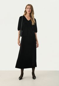 Schwarzes Samtkleid mit V-Ausschnitt, kurzen Puffärmeln und einem fließenden Rock. Kombiniert mit einer strukturierten schwarzen Tasche und spitzen Ballerinas.