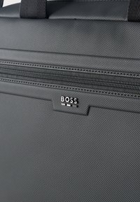 Μπροστινή όψη μαύρης υφασμάτινης τσάντας με οριζόντια φερμουάρ και ασημένιο λογότυπο BOSS κάτω από το τράβηγμα  του φερμουάρ.