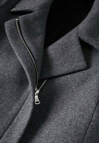 Abrigo de lana gris oscuro con un cierre de plata asimétrico parcialmente desabrochado cerca de las amplias solapas y el cuello.