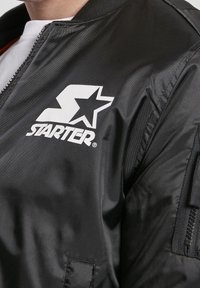 Giacca bomber nera con finitura lucida, collo a coste e un logo bianco prominente con una stella. Include un dettagliato taschino laterale con zip.