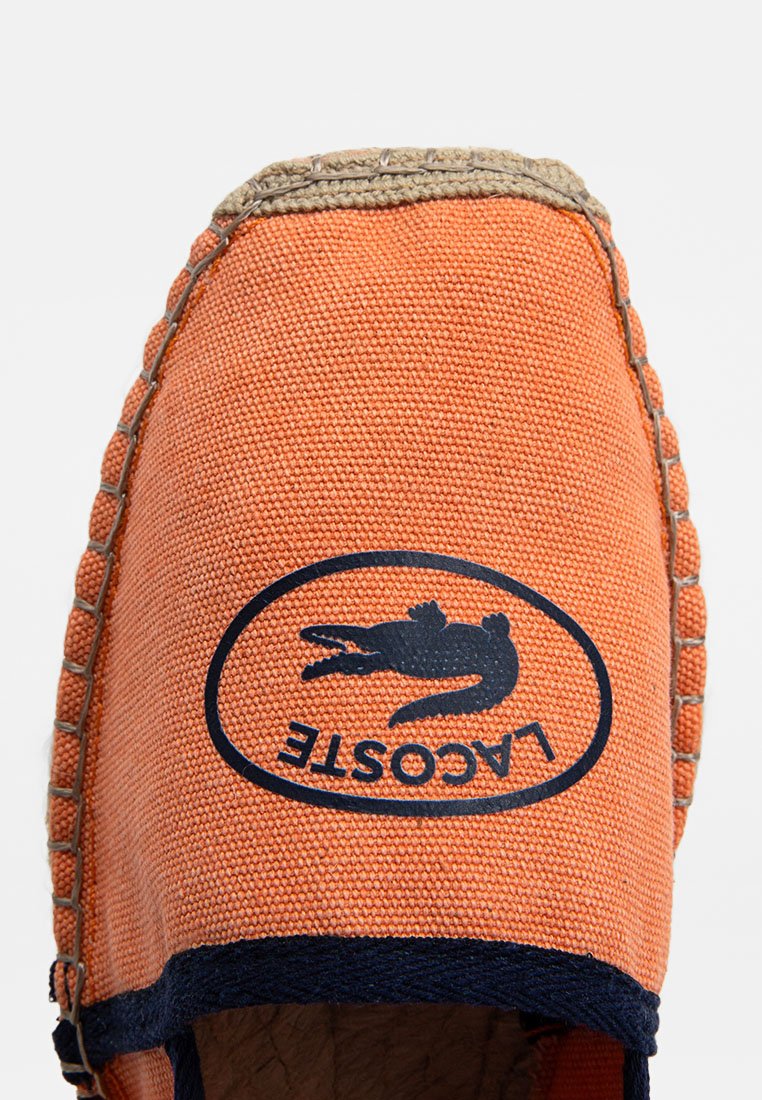 Espadrillas in tela arancione con una patch blu recante un design di coccodrillo, rifinite con trim in juta cucito e una punta arrotondata.