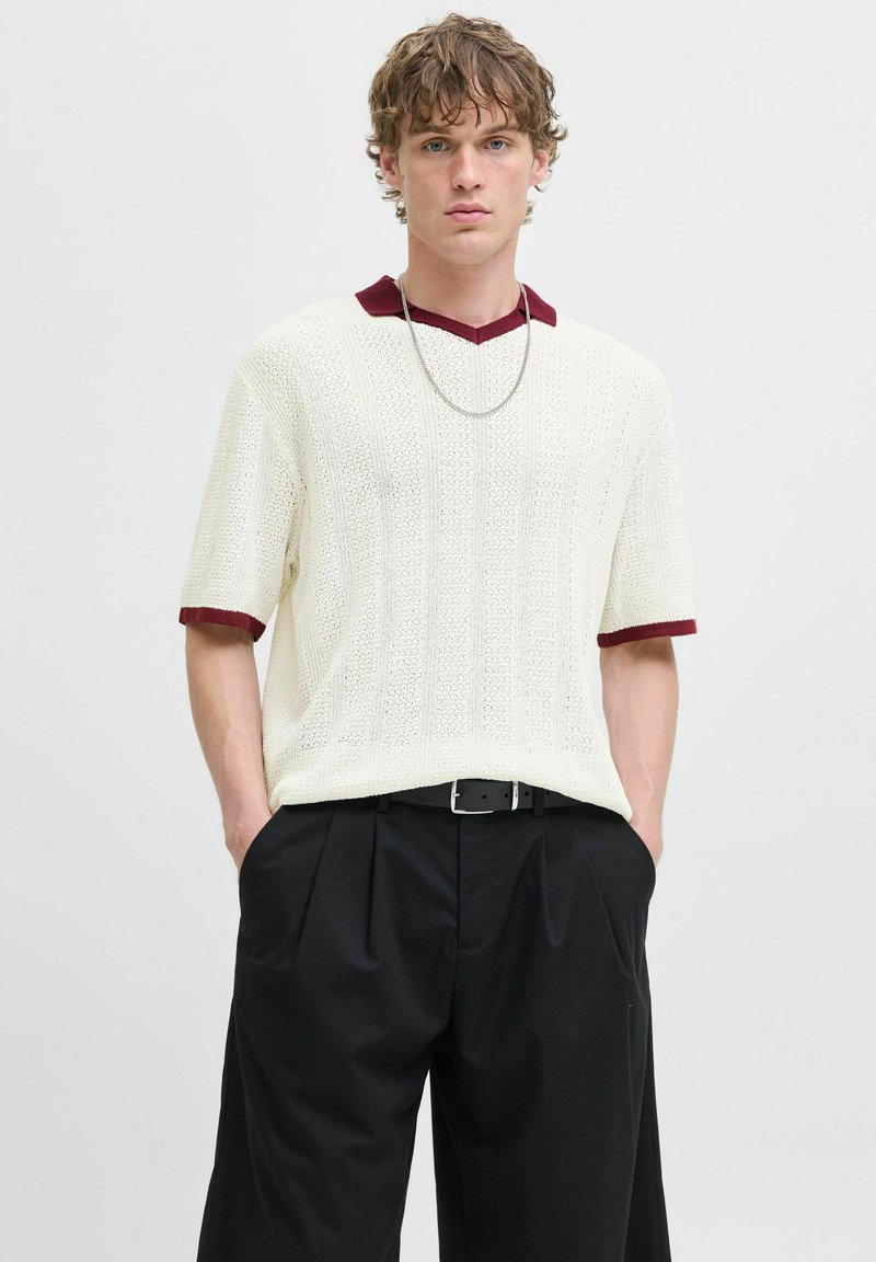 Hvid strikket kortærmet sweater med bordeaux farvet krave og manchetter, prydet med en struktureret søm. Sammen med sorte højtaljede bukser og et bælte.
