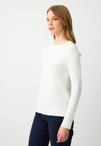 Maglione bianco lavorato a maglia con collo rotondo, maniche lunghe e polsini a coste. Tessuto morbido con una forma aderente, abbinato a pantaloni scuri.