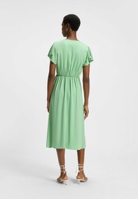 Vihreä, polvipituinen mekko, jossa on lyhyet kelluvat hihat ja elastinen vyötärö. Sujuva, virtaava kangas. Yhdistetty hihna-tyylisiin beigeihin sandaaleihin.