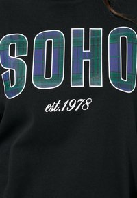 Camiseta de algodón negra con un gran texto blanco "SOHO" que presenta un patrón de cuadros verde y púrpura, y "est.1978" en una fuente blanca pequeña debajo.