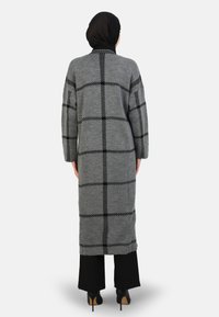 Lunga cardigan grigia lavorata a maglia con un motivo a scacchi neri, maniche larghe e spacco sui lati, abbinata a pantaloni neri e tacchi alti.