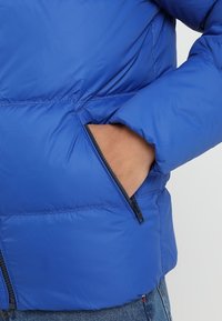 Blauwe pufferjack met een gladde, glanzende textuur. Voorzien van een ritszak en een gewatteerd design voor isolatie.