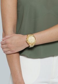 Reloj de pulsera cronógrafo dorado con una esfera redonda, marcadores de números romanos, correa de eslabones metálicos y caja texturizada. Lleva puesto en la muñeca sobre una blusa verde.