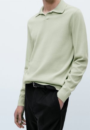 Homme portant un polo en maille vert clair à manches longues rentré dans un pantalon noir avec une ceinture noire, une main dans la poche.