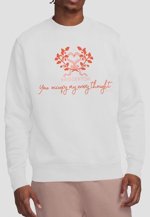 Mann trägt weißen Pullover mit rosa Herz- und Zweigdesign und dem Text „BRIDGERTON You occupy my every thought“ auf der Vorderseite.