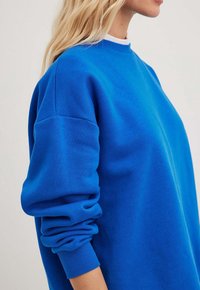 Sweatshirt bleu avec un col rond, des manches longues et des poignets côtelés. Le tissu semble doux avec une texture lisse et une coupe décontractée.