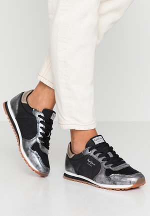 Persona con pantalones blancos y zapatillas Pepe Jeans negras y plateadas, de pie sobre un suelo gris claro.