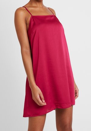 Femme portant une robe en satin rose-rouge, sans manches, ample, avec de fines bretelles spaghetti, debout devant un fond clair uni.