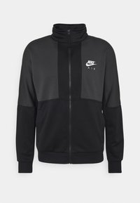Svart zip-up jacka i slät tyg med hög krage, som har en kontrasterande grå panel och en vit Nike Air-logga på bröstet.