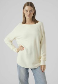 Cremefarbener Langarmpullover mit weitem Ausschnitt, weichem Material und lässiger Passform. Kombiniert mit hellblauen, locker sitzenden Jeans.