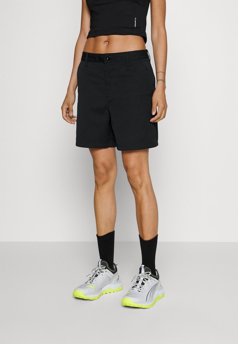 Person iført sorte knælange shorts, sorte mellemkabets sokker, hvide og neongule sneakers samt en sort kort top mod en ensfarvet baggrund.