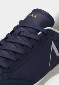 Αθλητικό παπούτσι σε ναυτικό μπλε με επάνω μέρος από υφή δέρματος, κορδόνια στον ίδιο χρωματισμό, λευκή επένδυση και χρυσό λογότυπο "ARKK" στη γλώσσα.