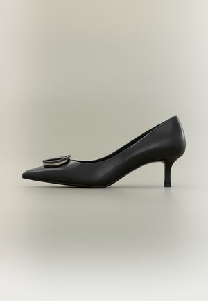 Escarpin en cuir noir à bout pointu et petit talon, avec une décoration boucle circulaire argentée sur le devant, sur fond beige uni.