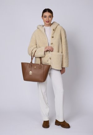 Chaqueta de piel sintética en crema con capucha y botones, combinada con un bolso de cuero marrón con un acento dorado, pantalones crema y zapatos color beige.