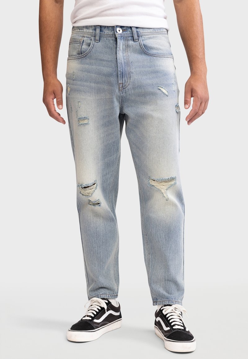 Lyseblå denimjeans med en afslappet pasform, slidte detaljer og falmet look. Kombineret med sorte og hvide sneakers, der viser en afslappet stil.