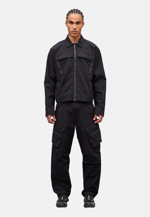 Uomo con capelli intrecciati che indossa una giacca nera con cerniera e tasche sul petto e pantaloni cargo neri con varie tasche con cerniera, in piedi.