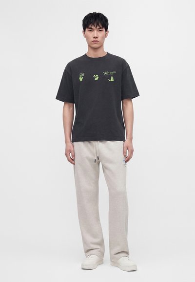 Νέος άνδρας στέκεται μπροστά φοράει μαύρο t-shirt Off-White με πράσινα γραφικά χεριών, μπεζ φόρμες και λευκά αθλητικά παπούτσια.
