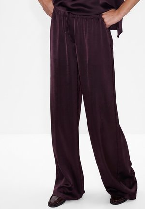 Pantaloni in raso viola scuro a gamba larga con vita elastica e coulisse, indossati con le mani in tasca e scarpe scure visibili.