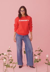 Jeune femme portant des lunettes violettes, un sweat-shirt rouge marqué "Amour" et un jean bleu, debout parmi des roses blanches dans des vases en verre sur un fond rose.