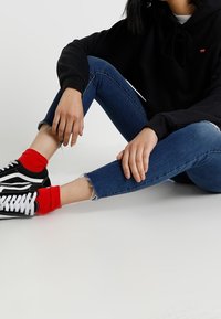 Sweat à capuche noir avec cordon de serrage, jean skinny bleu aux ourlets effilochés, chaussettes rouges, et baskets noires avec détails blancs. Tenue décontractée, en position assise.