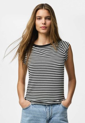 Ung kvinde med langt lyst brunt hår iført en sort-hvid stribet ærmeløs top og lyseblå denimjeans med hænderne i lommerne.