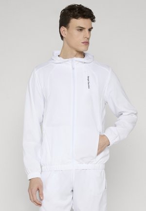 Mann iført en hvit hettejakke med glidelås og matchende bukser, med hånden i lommen, ser til siden mot en ensfarget bakgrunn.