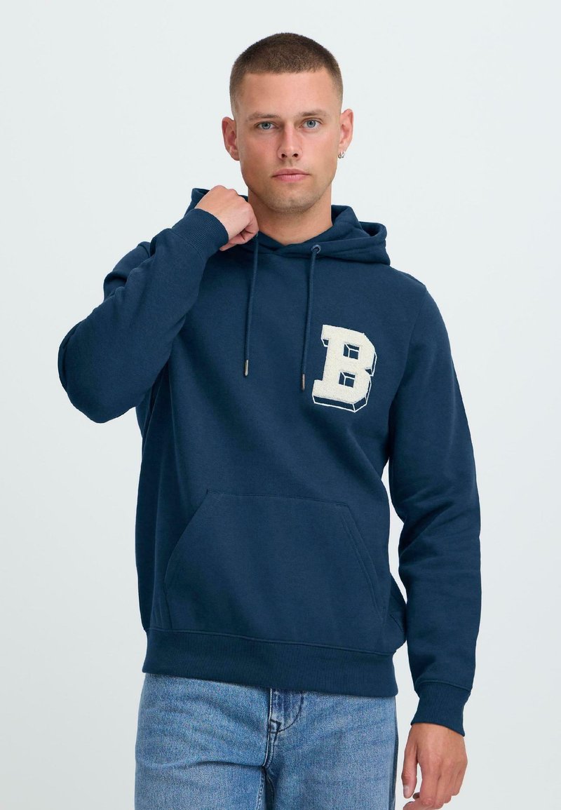 Jeune homme aux cheveux courts portant un hoodie bleu marine avec une grande lettre B blanche sur la poitrine et un jean bleu clair, se tenant contre un fond uni.