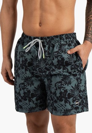 Homme portant un short de bain vert foncé avec un imprimé feuille tropicale noire, un cordon blanc et un petit logo circulaire sur la jambe gauche.
