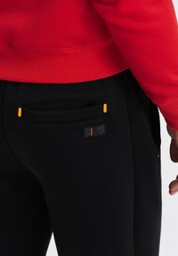 Sorte joggere med ribbet tekstur, med en orange detalje og logo-patch, parret med en rød sweater. Fokus på lommer og syninger.