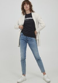 Helle Creme-Cardigan über einem schwarzen Grafik-T-Shirt, kombiniert mit blauen Skinny-Jeans und weißen Sneakers. Einfaches Design ohne zusätzliche Muster.