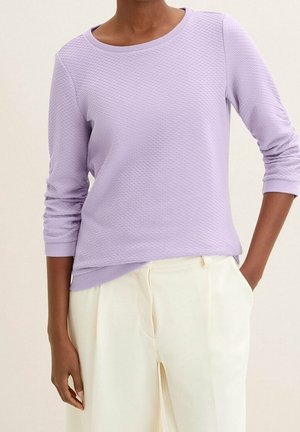 Frau trägt ein strukturiertes, lavendelfarbenes langärmeliges Oberteil, das in hoch taillierte, cremefarbene Hosen gesteckt ist, eine Hand in der Tasche, vor neutralem Hintergrund.