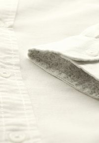 Witte textuur blouse met een knoopsluiting; de kraag onthult een subtiele grijze bladerenpatroon voering. Gemaakt van zachte katoen.