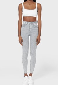 Jeans skinny de tiro alto en gris claro con una textura suave, cinco bolsillos y cierre de botón. Combinados con un top corto blanco y zapatillas blancas.
