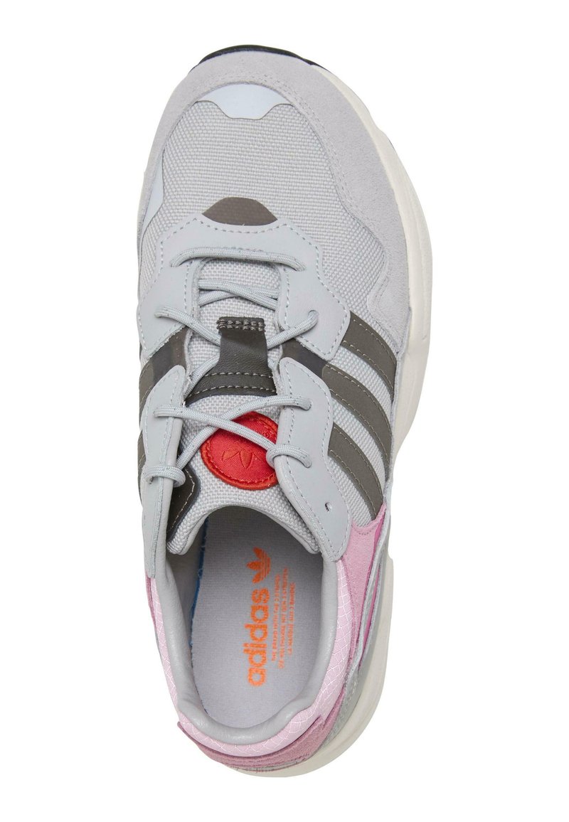 Scarpa sportiva grigia con pannelli in suede e mesh, caratterizzata da tre strisce grigie, dettagli rosa e un logo rosso sulla linguetta.